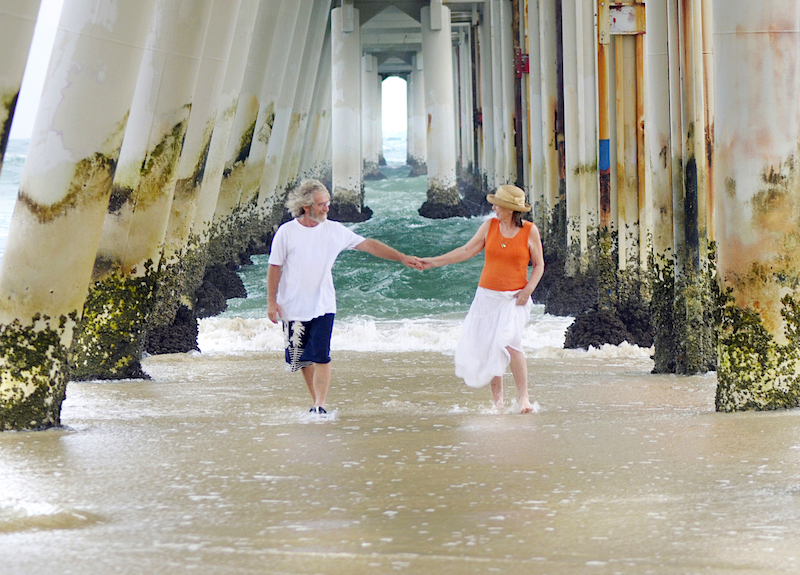 Couple holding hands and walking on a beach underneath a boardwalk