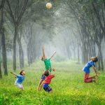 four children play with a ball outdoors, surrounded by trees