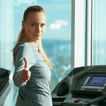 a blonde woman on a treadmill turns and gives a thumbs-up to the viewer