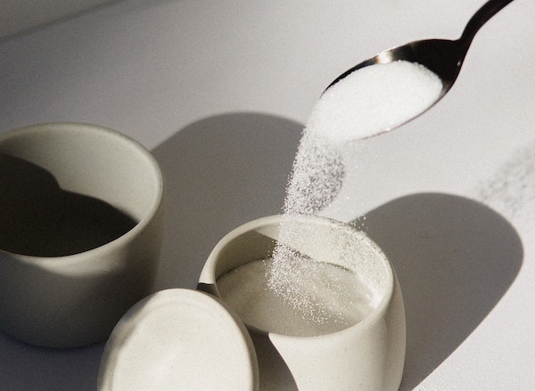 A white glass container of sugar with a spoon spilling sugar into it above