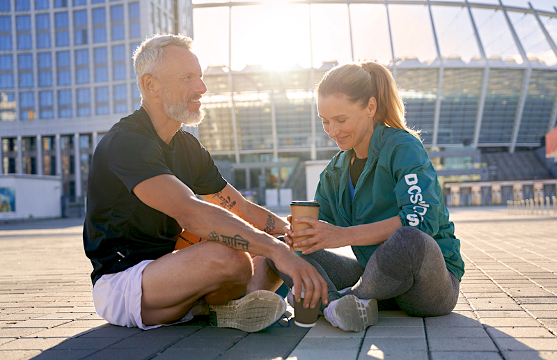An older couple sitting on a street in athletic wear. They are both holding cups of coffee.
